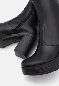 Bottes à la cheville en cuir noir avec talons carrés côtelés et épais, et fermetures éclair latérales, vues en gros plan sous un angle.