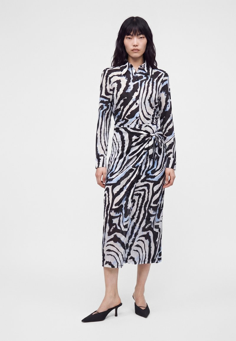 Γυναίκα που φοράει μακρυμάνικο, κουμπωτό φόρεμα με μαύρες, λευκές και μπλε αφηρημένες ζέβρες ρίγες, σε συνδυασμό με μαύρες μυτερές γόβες.