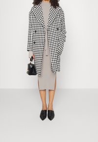 Manteau surdimensionné à motif pied-de-poule noir et blanc, robe grise côtelée, sac à main noir et chaussures pointues noires. Textures lisses et structurées.