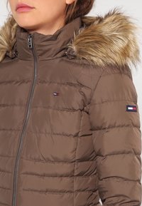 Brązowa kurtka puchowa z kapturem wykończonym futrem, z zapięciem na zamek błyskawiczny z przodu oraz pionowymi detalami szwów. Łatki z logo na rękawach. Gładka tekstura.