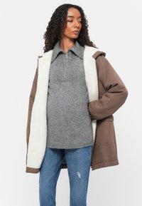 Hnědý vnější kabát s bílou umělou kožešinou uvnitř, šedý svetr na zip pod ním a modré oprané džíny. Model stojí na bílém pozadí.