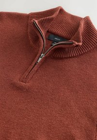 Ruosteenvärinen keskipainoinen neulepaita, jossa on resorireunainen korkea kaulus ja puoliksi auki oleva metallivetoketju, joka paljastaa kokolapun.