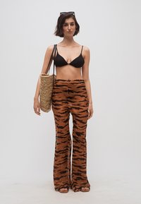 Γυναίκα που φοράει μαύρο μπικίνι τοπ, καφέ παντελόνι με ρίγες τίγρης, σανδάλια, κρατώντας πλεκτή τσάντα ώμου, με γυαλιά ηλίου στο κεφάλι, στέκεται μπροστά σε απλό φόντο.