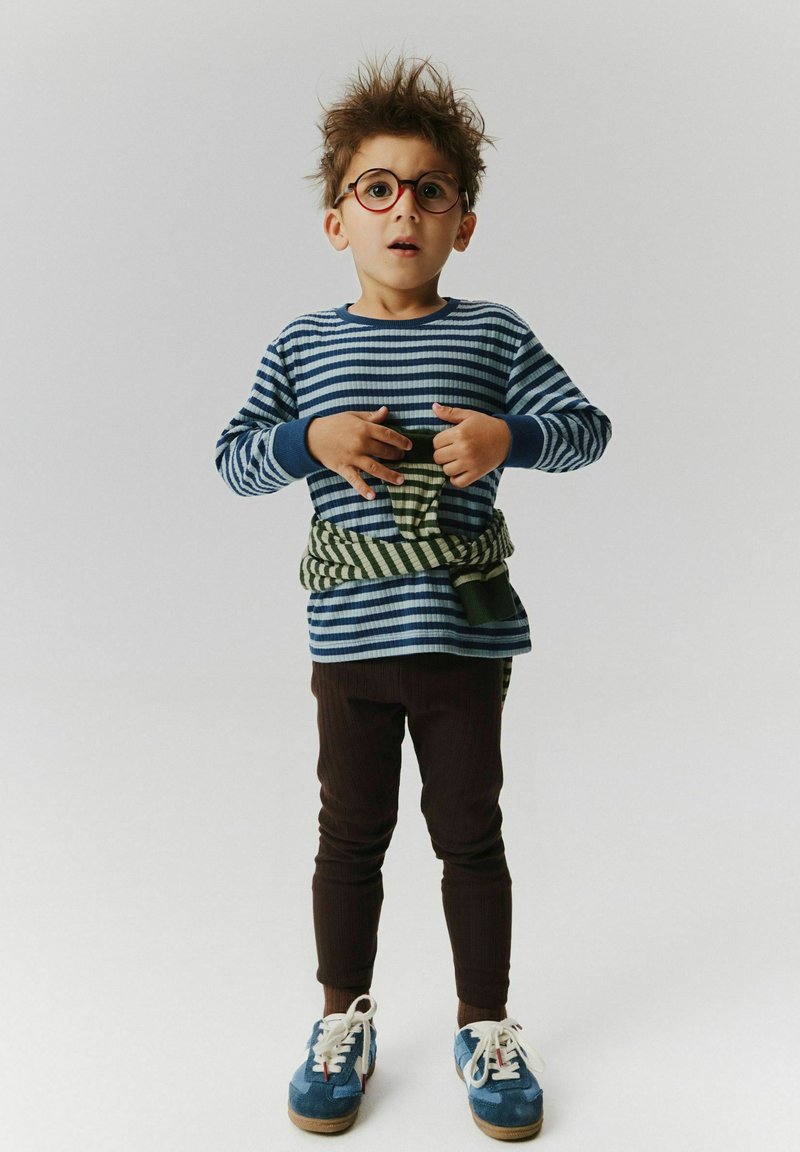 Jeune garçon aux cheveux en bataille et portant des lunettes, habillé d'une chemise rayée, d'un pantalon et de baskets, tenant un pull rayé vert autour de sa taille.