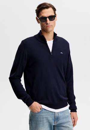 Mees, kes kannab musti päikeseprille, tumesinist lukuga kampsunit valge särke peal ja helesiniseid teksaseid, seisab ühe käe taskus.