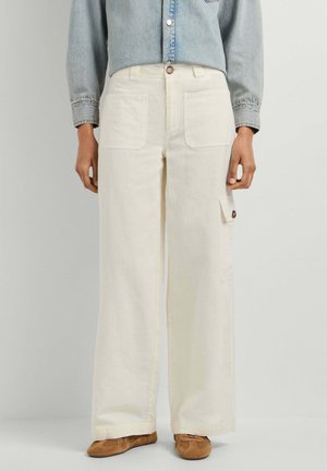Persona que lleva pantalones anchos de talle alto en color crema con bolsillos parche delanteros y laterales, combinados con zapatos marrones y una camisa vaquera.
