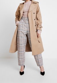 Beżowy płaszcz trench z podwójnym zapięciem i talią ze sznurkiem, w połączeniu z kraciastymi spodniami w jasnoszarym, czarnym i żółtym odcieniu.
