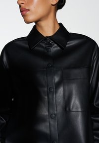 Camicia in pelle nera con colletto a punta, chiusura con bottoni e una tasca sul petto. Texture liscia con dettagli cuciti.