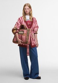 Übergroßer Cardigan in Pink und Rot mit Muster und weiten Ärmeln, kombiniert mit blauen weiten Jeans und einer großen mehrfarbigen gesteppte Handtasche.