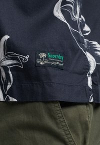 Camisa de manga corta azul marino con patrones florales blancos, con una etiqueta tejida de Superdry, combinada con pantalones oscuros de tela color verde oliva.