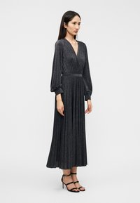 Μακρύ μαύρο φόρεμα με βαθύ V-λαιμόκοψη, πλισέ φούστα και μακριά μανίκια, από υφή υφάσματος που αστράφτει. Συνδυάζεται με μαύρα τακούνια με λουράκια.