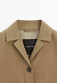 Khaki bunda s výstužovým límcem, s jedním knoflíkem na zapínání a viditelným švem. Na vnitřním štítku je označena "Massimo Dutti".