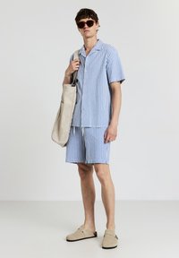 Gestreiftes Kurzarmhemd und Shorts-Set in Blau und Weiß, aus leichtem Material gefertigt. Enthält eine beigefarbene Schultertasche und Sandalen.