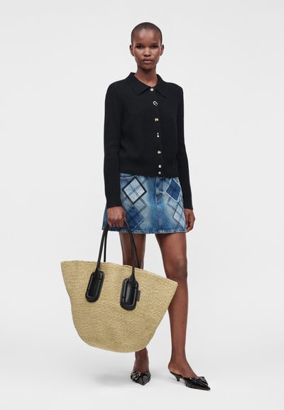 Femme portant un cardigan noir et une jupe en jean bleue à motifs, tenant un grand sac tissé beige avec des poignées noires, debout sur fond blanc.