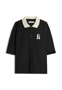Zwarte polo met korte mouwen en een crèmewitte kraag en knopen, voorzien van een geborduurde letter "K" op de linkerkant van de borst.
