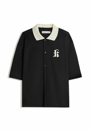 Polo shirt nero a maniche corte con colletto e bottoni color crema, dotato di una lettera "K" ricamata nell'area sinistra del petto.