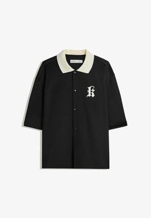 Polo shirt nero a maniche corte con colletto e bottoni color crema, dotato di una lettera "K" ricamata nell'area sinistra del petto.