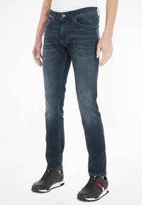 Jeans de mezclilla azul oscuro con corte ajustado, que muestran un diseño clásico de cinco bolsillos y un sutil detalle de costura. Combinados con zapatillas deportivas negras.