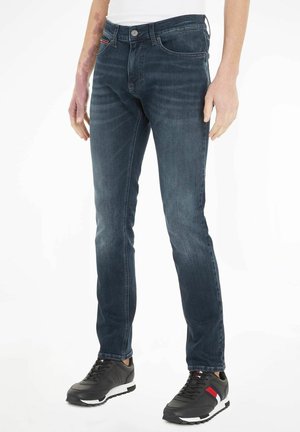 Jeans de mezclilla azul oscuro con corte ajustado, que muestran un diseño clásico de cinco bolsillos y un sutil detalle de costura. Combinados con zapatillas deportivas negras.
