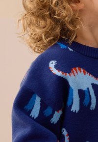 Tmavomodrý sveter s motívmi dinosaurov v červenej, modrej a bielej farbe. Mäkká textúra, okrúhly výstrih, viditeľné rebrované manžety a lem.