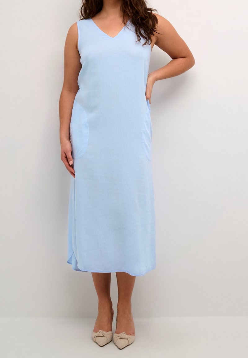 Femme portant une robe midi bleu clair sans manches avec des poches latérales et des chaussures beige à bouts pointus, debout devant un fond uni.