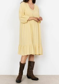 Robe jaune à col en V avec un motif géométrique, manches 3/4 et ourlet froncé. Portée avec des bottes marron jusqu'aux genoux. Texture lisse.