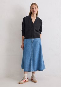 Cardigan noir à col en V avec des boutons, associé à une longue jupe en denim bleu clair. Le modèle porte des baskets beiges avec des rayures rouges et des chaussettes claires.