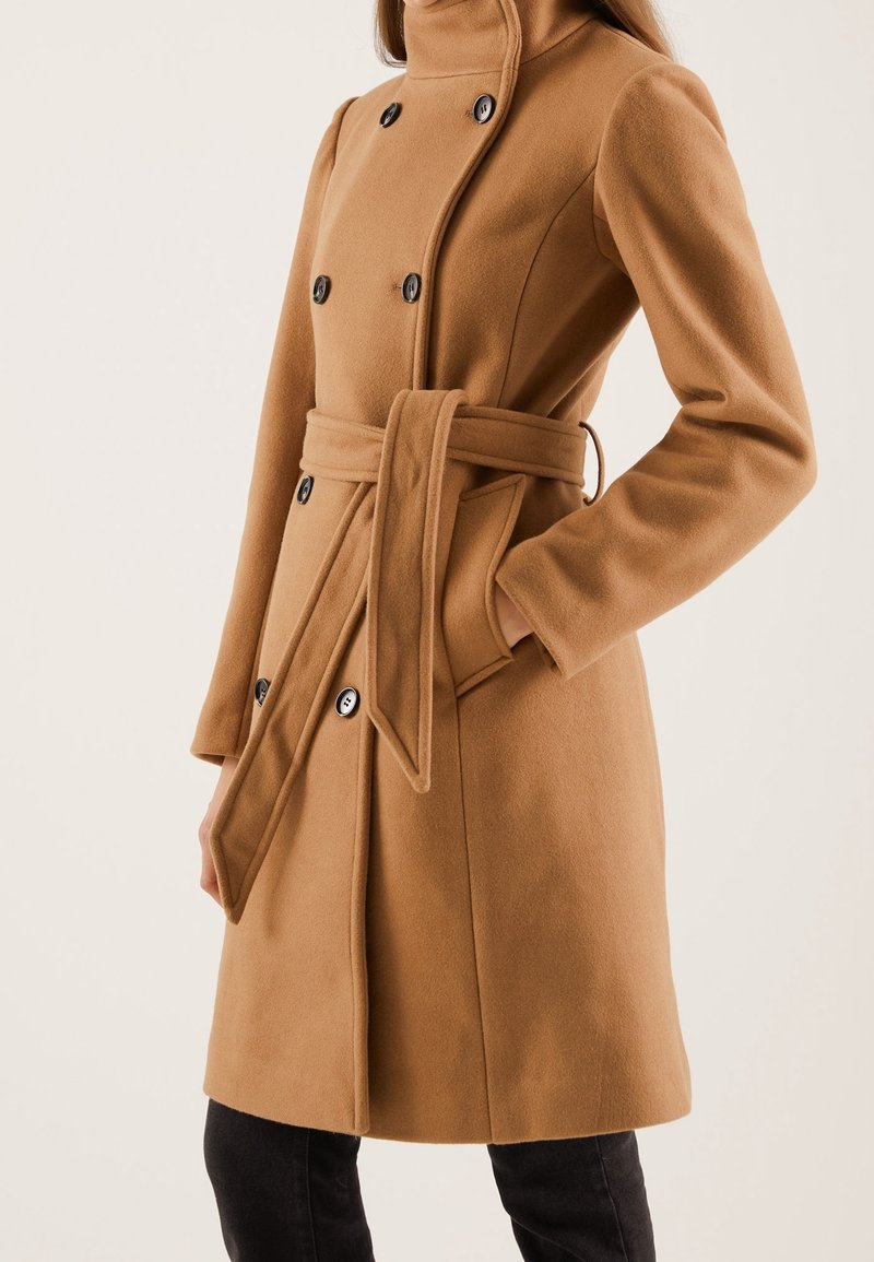 Kamelivillasekoituksesta valmistettu takki, jossa korkea kaulus, kaksiriviset napit, solmittava vyö vyötäröllä ja sivutaskut. Sileä pinta ja räätälöity istuvuus.