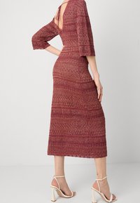 Robe en tricot bordeaux avec un motif texturé, manches trois-quarts, dos ouvert avec un détail de nœud, et longueur au mollet. Portée avec des talons blancs à lanières.