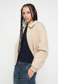 Casaco bomber bege em mistura de lã com gola, bolsos frontais e ferragens douradas, combinado com um suéter canelado azul-marinho e jeans de ganga azul.