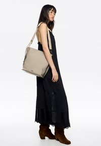 Beżowa torba na ramię o gładkiej teksturze, prostokątnym kształcie i regulowanym pasku. Delikatne logo z przodu. Modelka ma na sobie czarną sukienkę.