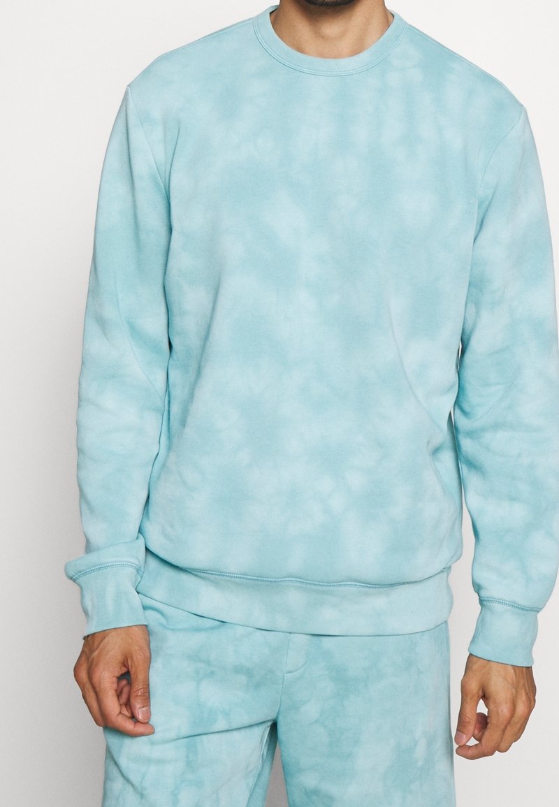 Homme portant un sweat-shirt à teinture sur tissu bleu clair et un pantalon de survêtement assorti, debout avec les mains détendues le long du corps devant un fond uni.