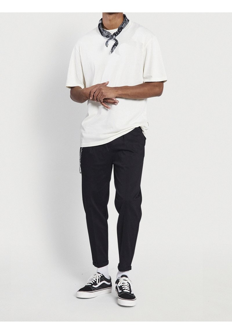 Homme portant un t-shirt blanc, un pantalon noir, des baskets noires et blanches, et un bandana sombre à motifs autour du cou, debout les mains jointes.