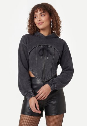 Kvinde iført en vasket sort cropped hoodie med korsetlignende frontlynlås og sorte lædershorts, stående med hænderne foldet.
