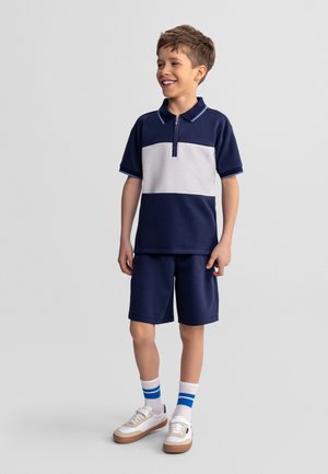 Garçon souriant portant un polo à manches courtes bleu marine et blanc, un short bleu marine, des chaussettes blanches avec des rayures bleues et des baskets blanches, debout devant un fond gris.