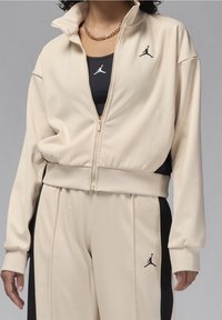 Chaqueta crema con cremallera y acentos negros, con un alto cuello y el logo de Jordan. Combinada con una camiseta sin mangas negra y pantalones a juego.