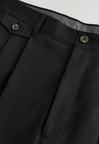 Pantalon noir plissé avec ceinture, passants de ceinture, poche latérale à rabat et deux boutons foncés sur la fermeture avant.