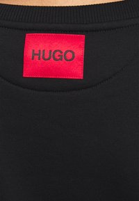 Camiseta de tela negra con textura acanalada y escote redondo. Presenta una etiqueta rectangular roja con "HUGO" en letras negras en la parte trasera.
