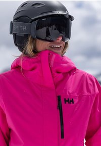 Helle pinkfarbene, wasserdichte Skijacke mit schwarzem Logo, ausgestattet mit einem hohen Kragen, einer Reißverschlusstasche und kombiniert mit einem schwarzen Helm und einer Skibrille.