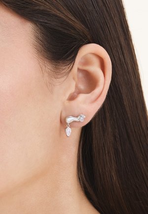 Silver earring with wavy design and two crystal stones worn on the left ear of a person with dark brown hair.