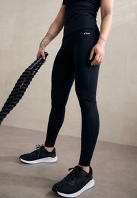 Leggings atletici neri con design senza cuciture e leggero lucido, abbinati a scarpe da allenamento nere con suola bianca e lacci testurizzati.