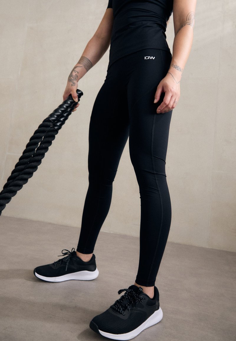 Leggings atletici neri con design senza cuciture e leggero lucido, abbinati a scarpe da allenamento nere con suola bianca e lacci testurizzati.