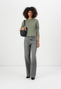 Olijfgroene geribde lange mouwen top, hoge taille grijze wijde jeans, zwarte handtas en puntige hakken, geplaatst tegen een effen achtergrond.