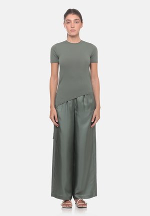 Donna in piedi frontalmente, indossa una t-shirt aderente verde oliva a maniche corte e pantaloni larghi in satin con sandali su sfondo semplice.
