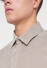 Camisa de suave color gris claro en tela de pana, con un cuello clásico, que presenta dos cierres de botón, textura de canalé vertical y un corte ligeramente relajado.