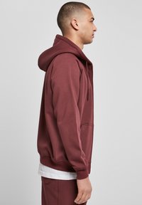 Miestenská marónová mikina vyrobená z mäkkej látky. Má klokanie vrecko, kapucňu so šnúrkou a rebrované manžety. Nosená cez biele tričko.