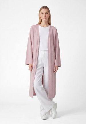 Kvinde stående iført en lang lyserød cardigan med åben front, hvid skjorte, brede hvide bukser og hvide sneakers mod en ensfarvet baggrund.