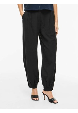 Nő laza fekete mandzsettás nadrágban, oldalzsebekkel és fekete nyitott orrú magas sarkú szandálban, fehér háttér előtt állva.