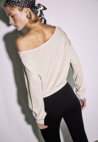 Ženska obračajoča se nazaj, oblečena v bež pulover brez ramen, črne hlače, ruto z belimi pikami in zlati obroči na ušesih, na enobarvnem ozadju.