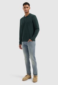 Donkergroene polo met lange mouwen en een gestructureerd patroon, met een knoopsluiting met twee knopen, gecombineerd met lichtblauwe spijkerbroek en beige suède schoenen.